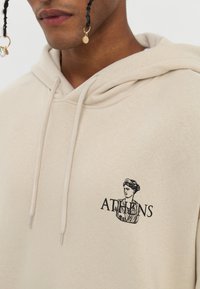 Beige hoodie in soft fabric featuring a graphic of a classical bust and "ATHENS" text in black on the chest. Drawstring hood details visible.