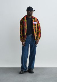 Veste en polaire à carreaux rouges et jaunes avec une fermeture éclair, t-shirt noir, jean bleu ample et casquette noire. Lunettes de soleil dorées et chaussures noires.