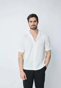 Chemise blanche à manches courtes, boutonnière avec un motif texturé, dotée d'un col à revers et de boutons bleu clair, associée à un pantalon noir.