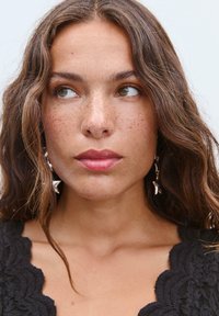 Primo piano di una persona con capelli castani ondulati, che indossa orecchini d'argento a forma di stelle e un indumento nero con texture e bordi ondulati.