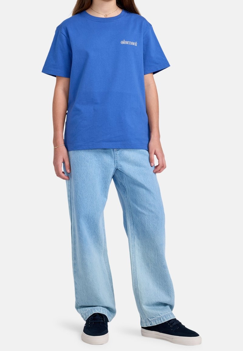 Personne portant un t-shirt bleu avec le logo Element, un jean bleu clair ample et des baskets noires à semelles blanches, debout devant un fond uni.