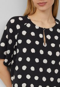 Schwarze Bluse mit weißen Punkten, runder Ausschnitt und Keyhole-Detail, lockere Passform, kurze Ärmel, strukturierter Stoff.