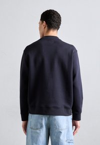 Felpa Navy con collo a giro e polsini a coste. Tessuto liscio, vestibilità rilassata. Presenta un piccolo etichetta sul retro del collo ed è abbinata a jeans azzurri.