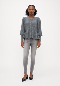 Grijze gebreide cardigan met een peplum taille, lange puffmouwen en knoopsluiting, gecombineerd met lichtgrijze skinny jeans en zwarte hakken.
