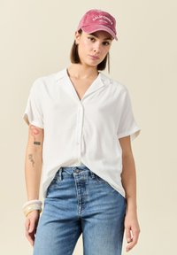 Camisa blanca de manga corta con botones y un patrón texturizado, combinada con jeans de mezclilla azules y una gorra rosa con "CALIFORNIA" en blanco.