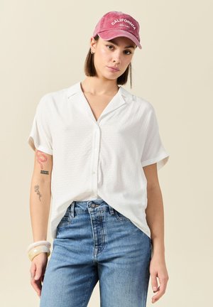 Camisa blanca de manga corta con botones y un patrón texturizado, combinada con jeans de mezclilla azules y una gorra rosa con "CALIFORNIA" en blanco.