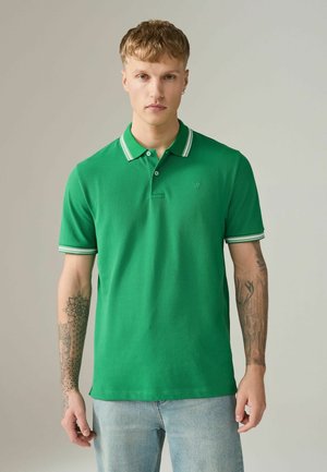 Giovane uomo con capelli biondi corti e tatuaggi su entrambe le braccia, indossa una polo verde e jeans azzurri, in piedi davanti a uno sfondo semplice.