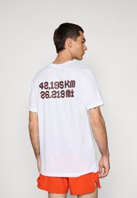 Man wearing white T-shirt with "42,195 km 26.219 mi" text on back and bright orange shorts, standing against plain background.
