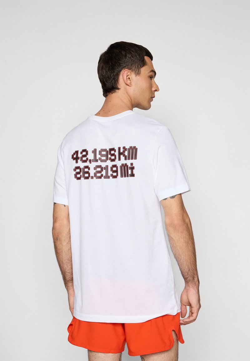 Man wearing white T-shirt with "42,195 km 26.219 mi" text on back and bright orange shorts, standing against plain background.