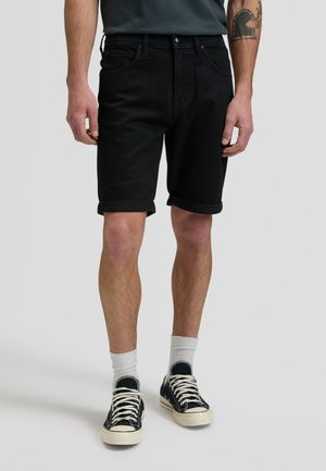 Mees, kes kannab musti, ülespoole rullitud teksapükse, valgeid keskmise pikkusega sokke ja musti madala säärega tossusid, seisab tavalise halli tausta ees.