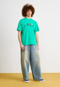 Green cotton t-shirt with black and multicolored "Valcom Cosmic Love Party" text, paired with loose-fitting blue denim jeans.