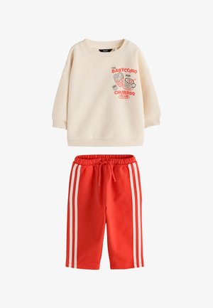 Lille barn iført cremefarvet sweatshirt med rød og sort tryk, der viser churros i en kegle og en kop mærket "The Babyccino and Churros Club."