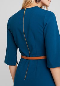 Teal klänning med bakdragkedja och brun bälte. Har en figurnära design, ärmskärning till armbågen och en mjuk tygstruktur.