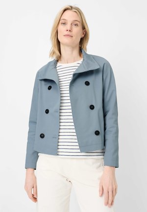 Woman wearing light blue double-breasted jacket over white and navy striped shirt and cream-colored pants against a plain background.