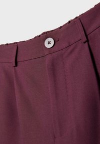 Detailný záber na pás bordeaux nohavíc s gombíkom, pútkom na opasok a elastickým chrbtom na jednofarebnom pozadí.