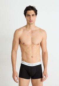 Boxer-briefs en coton noir avec une taille logo TOMMY HILFIGER blanche. Modèle présenté avec un torse nu sur fond blanc.