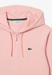 Rosa zip-up hoodie i mjukt tyg, med en grön broderad krokodil-logotyp på bröstet och dragsnören i huvan.