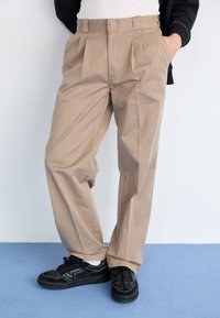 Béžové kalhoty s plisováním a vysokým pasem, vyrobené z hladké látky; nošené s černými teniskami s bílými akcenty a texturovanými detaily.