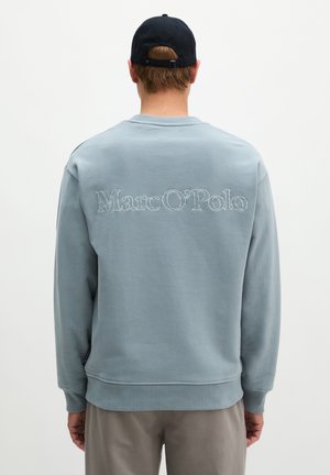 Mężczyzna w jasnoniebieskiej bluzie z haftowanym na plecach napisem "Marc O'Polo" oraz czarną czapką, zwrócony tyłem do kamery.