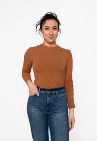 Frau mit dunklen Haaren, die ein braunes, geripptes Langarmshirt und blaue Jeans mit einem schwarzen Gürtel trägt, steht mit einer Hand in der Tasche vor einem weißen Hintergrund.