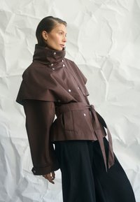 Nő barna, övvel díszített dzsekit visel, magas gallérral és széles ujjal, fekete, bő szárú nadrággal párosítva, gyűrt fehér háttér előtt pózol.