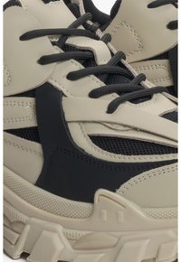 Beżowe i czarne sneakersy z teksturowaną powierzchnią, panelami z siateczki, grubą podeszwą oraz czarnymi sznurówkami. Wyróżniają się warstwowym designem i detalami w postaci szwów.