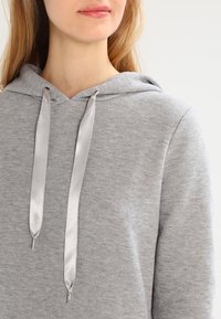 Detailní záběr na osobu v světle šedé mikině s širokými, lesklými saténovými šňůrkami, na jednobarevném světlém pozadí.