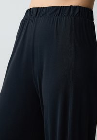 Primo piano della vita e delle gambe di una persona che indossa pantaloni neri con elastico in vita, realizzati in un tessuto liscio e morbido, su uno sfondo neutro.