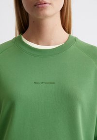 Grön sweatshirt med rund halsringning och långa raglansärmar. Har en mjuk textur och en liten präglad logotyp i en mörkare nyans.