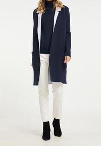 Langer marineblauer Cardigan aus weichem Stoff mit weißem Akzent-Kapuze, ausgestattet mit zwei Fronttaschen und offenem Design. Kombiniert mit weißen Hosen und schwarzen Stiefeln.