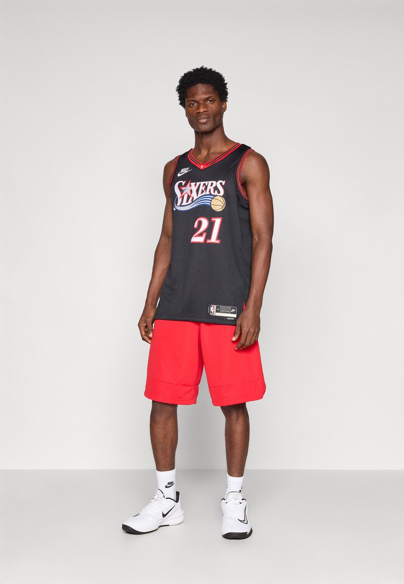 Black basketball jersey with red trim and "SIXERS" print; paired with red shorts and white sneakers. Model stands against a neutral background.