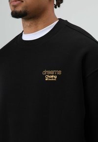 Zwarte sweatshirt met geribbelde halslijn en gele geborduurde tekst: "dreams Chasing NOT deadlines." Zachte textuur, comfortabele pasvorm.