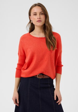 Donna che indossa un maglione arancione ampio con maniche tre quarti e una gonna di jeans scura con cintura marrone, in piedi davanti a uno sfondo semplice.