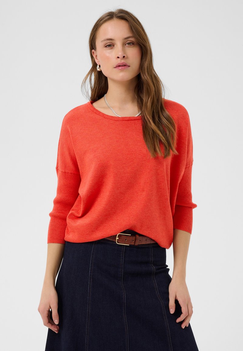 Femme portant un pull orange ample à manches trois-quarts et une jupe en jean foncé avec une ceinture marron, debout devant un fond uni.