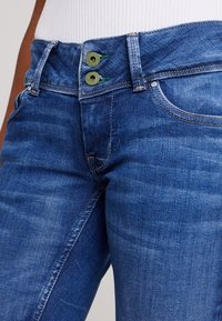 Jeans i denim i en medelblå färg med högmidjad design, två gröna knappdetaljer och subtil textur.