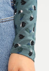 Hihna vihreästä paidasta, jossa on musta, valkoinen ja sininen abstract-kukkakuvio, lepäämään sinisten farkkujen taskulle.