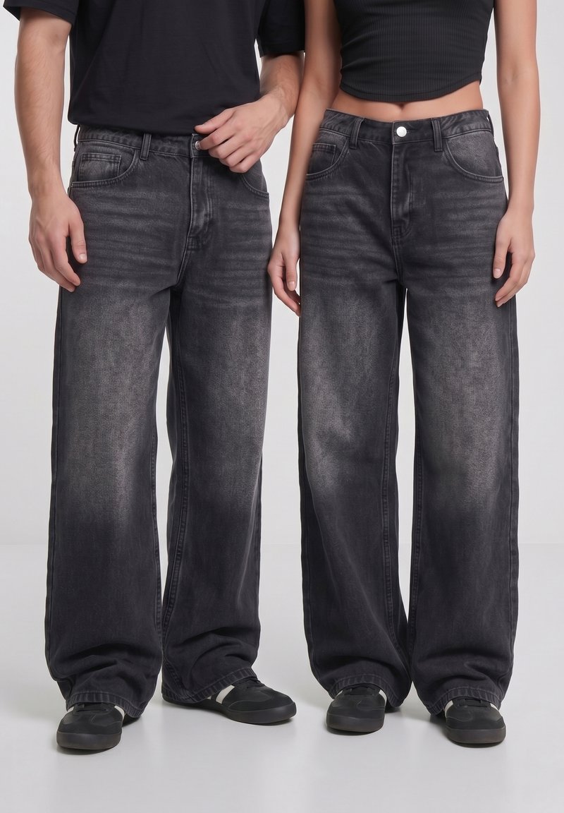 To personer iført sorte falmede jeans med vide ben og sorte sneakers, stående side om side mod en ensfarvet baggrund.