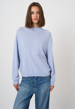 Jonge vrouw met schouderlang bruin haar, die een lichtblauwe trui en blauwe jeans draagt, staand voor een effen witte achtergrond.