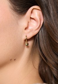 Złote kolczyki w kształcie kół z małym, wiszącym serduszkiem. Powierzchnia jest gładka i lustrzana, prezentując wypolerowane metaliczne wykończenie.