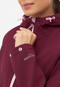 Burgundowa wodoodporna kurtka z kapturem na sznurku, kontrastowymi jasnoróżowymi akcentami oraz srebrnym okuciem z logo na rękawie.