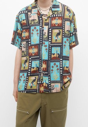 Chemise à manches courtes boutonnée avec des motifs colorés encadrant des oiseaux, fleurs et formes géométriques, portée avec un pantalon vert olive doté de poches zippées.