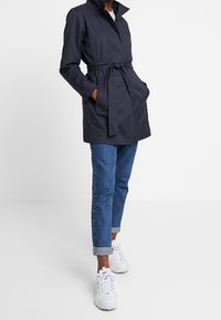 Trench coat leggero color blu navy con vita alta e cinturino, tasche con patta e colletto. Indossato sopra jeans azzurri arrotolati e sneakers bianche.