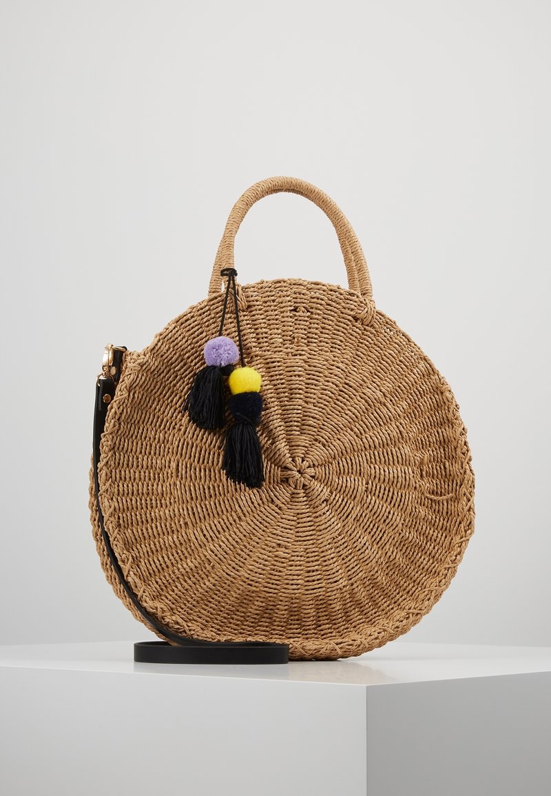 Sac à main rond en paille tressée avec poignée supérieure et bandoulière noire, décoré de pompons violets et jaunes ainsi que de glands noirs, exposé sur une surface blanche.