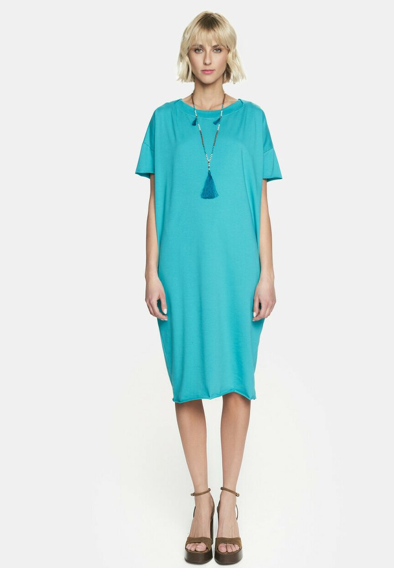 Turkusowa sukienka o krótkim rękawie, luźnym kroju, z okrągłym dekoltem i naszyjnikiem z frędzlami. Gładki materiał i długość do kolan.