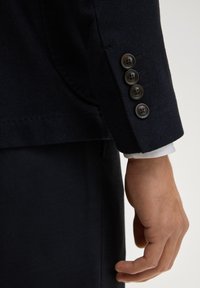 Manche de blazer bleu marine avec quatre boutons foncés, tissu texturé, avec un poignet de chemise blanche visible en dessous. Main reposant à côté.