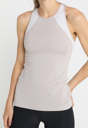 Femme portant un débardeur de sport gris clair avec des panneaux en mesh et un legging noir, debout avec une main sur la hanche.