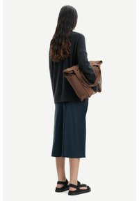 Maglione nero a maglia, gonna blu navy fino al ginocchio, sandali neri e una borsa marrone con tracolle, visto da dietro su uno sfondo chiaro.
