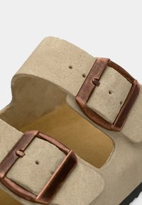 Sandale en daim beige avec boucles réglables en ton cuivre, tige texturée et semelle intérieure rembourrée de couleur tan, avec un design à bout ouvert.