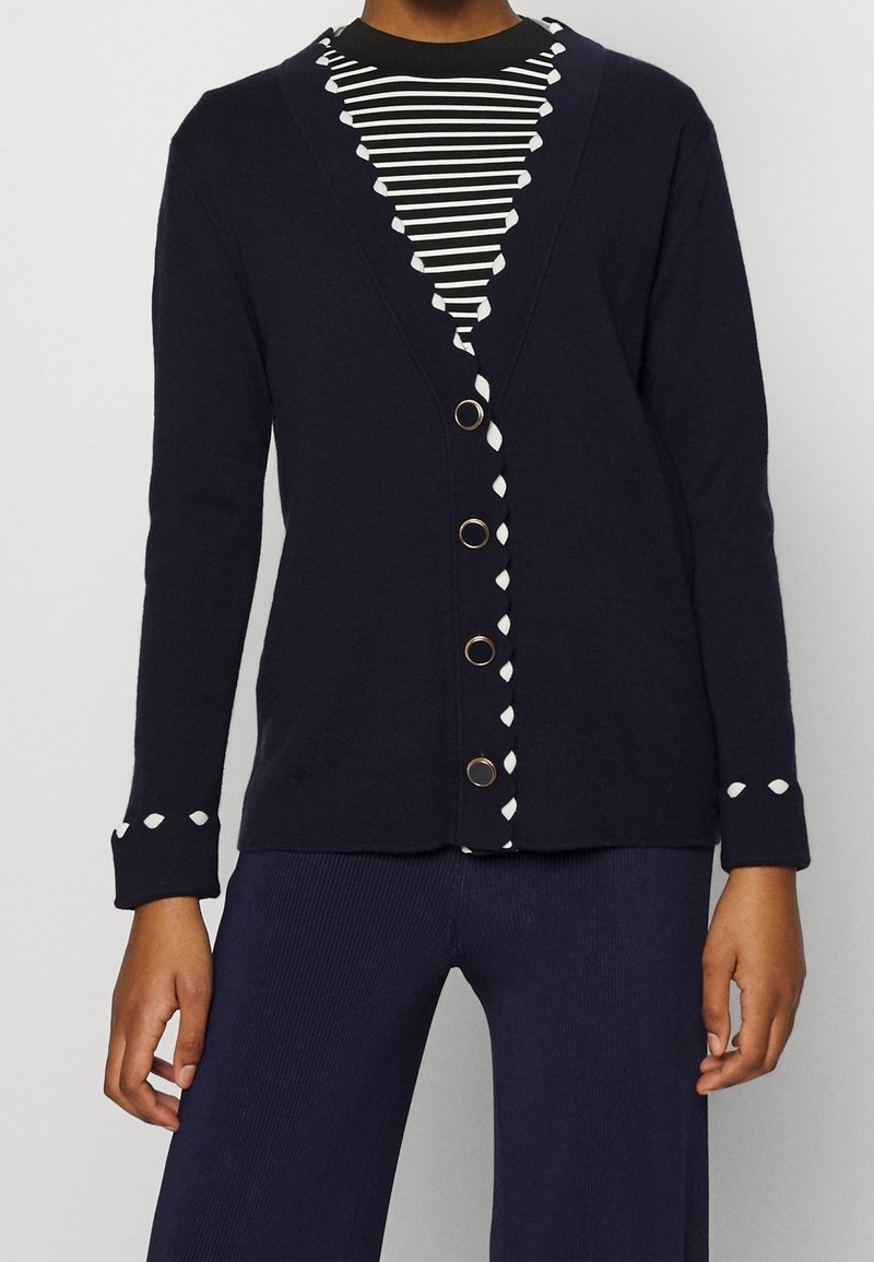 Mörkblå kofta med djup V-ringning, vit kant längs kanten, runda guldknappar och långa ärmar, lagd över en randig svart-och-vit topp.