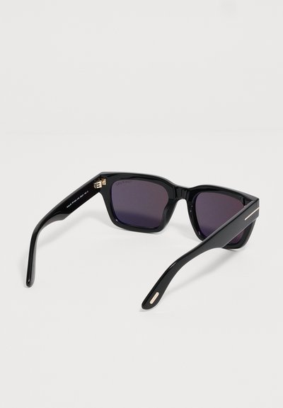 Lunettes de soleil rectangulaires noires avec des verres teintés foncés et des branches légèrement courbées, reposant ouvertes sur une surface blanche.